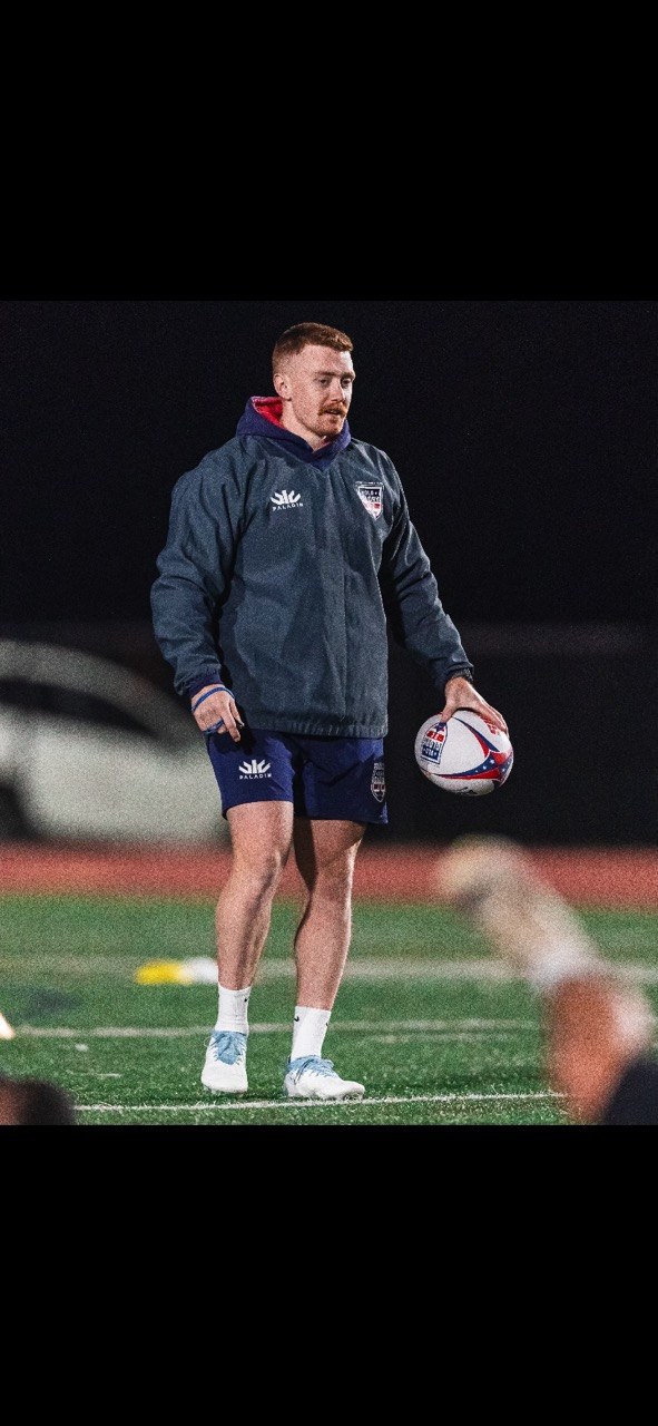 Mathew Skyner holding a rugby ball on a floodlit field at night